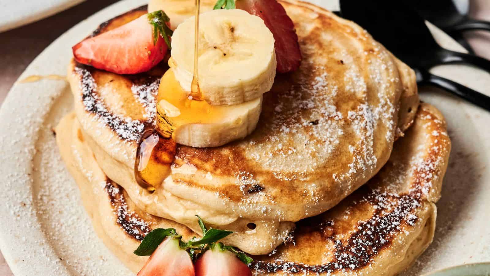 A stack of pancakes topped with sliced bananas, strawberries, and syrup on a plate, accompanied by black utensils.
