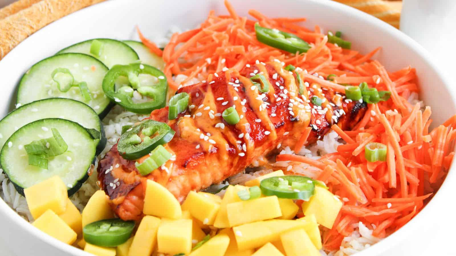 A bowl with grilled salmon on rice, topped with sesame seeds, surrounded by sliced cucumbers, shredded carrots, jalape&ntilde;os, mango cubes, and chopped green onions.