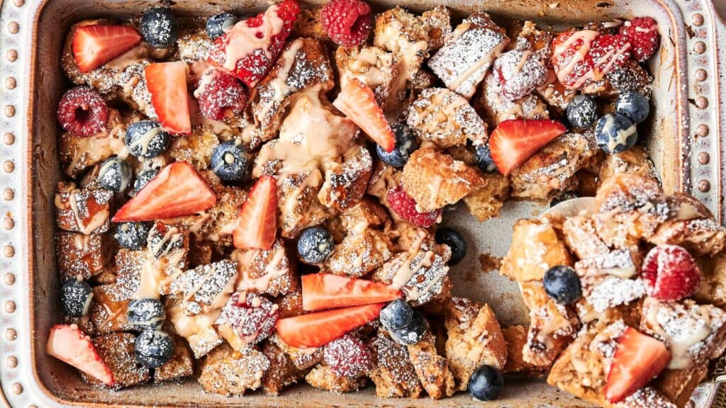 A baked casserole topped with powdered sugar, strawberries, raspberries, blueberries, and a drizzle of sauce in a rectangular dish.