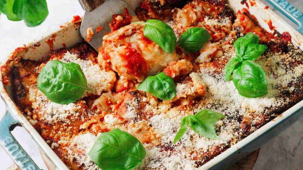 A baked dish of lasagna topped with tomato sauce, grated cheese, and fresh basil leaves, with a serving spoon taking a portion from the corner.