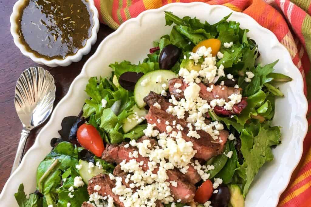Salad with mixed greens, sliced steak, cherry tomatoes, cucumbers, and crumbled cheese on a white plate. A bowl of dressing and a spoon are beside it on a wooden surface.