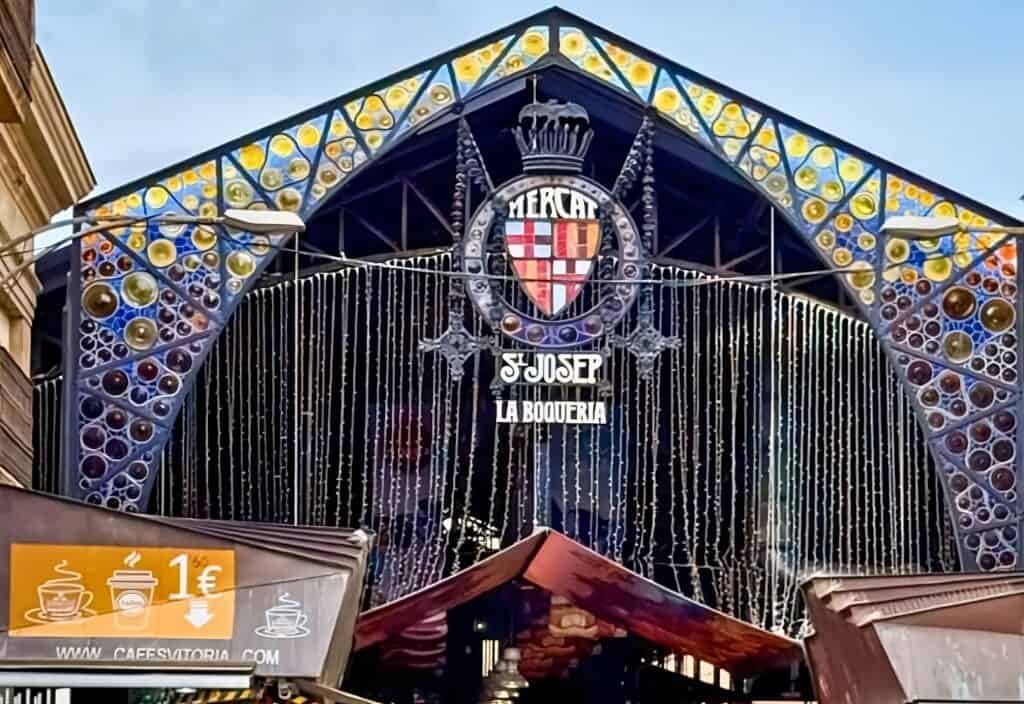 At the entrance of Mercat de Sant Josep de la Boqueria, known as a haven in Barcelona for foodies, decorative stained glass and outdoor stalls entice visitors, with a prominent coat of arms gracefully poised above.