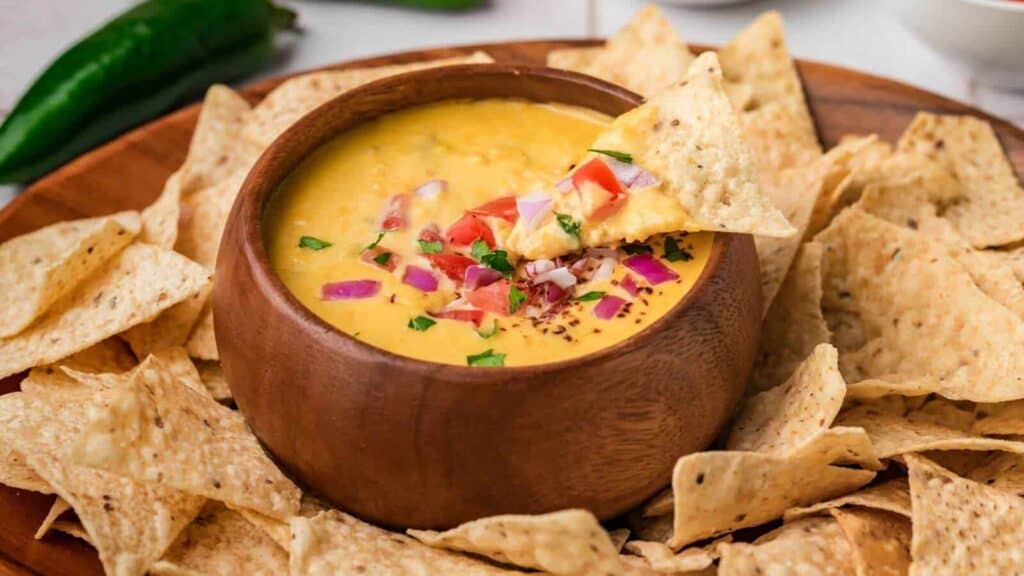 A wooden bowl of cheese dip topped with diced tomatoes and onions, surrounded by tortilla chips on a wooden platter.