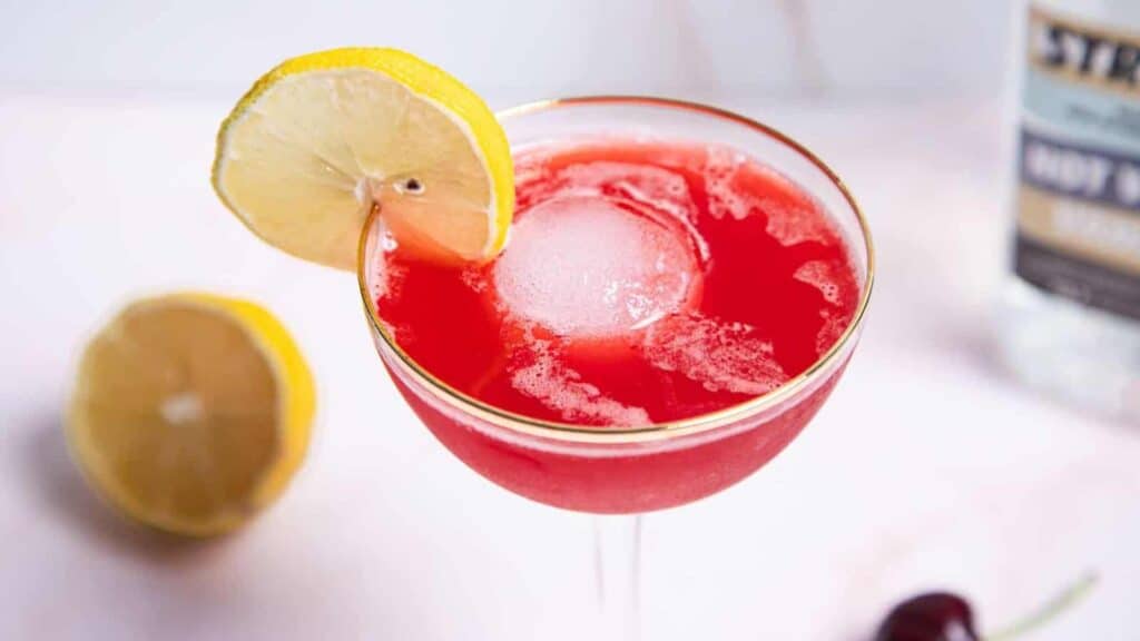 Red cocktail in a stemmed glass with a large ice sphere, garnished with a lemon slice. Half a lemon and a cherry rest nearby.
