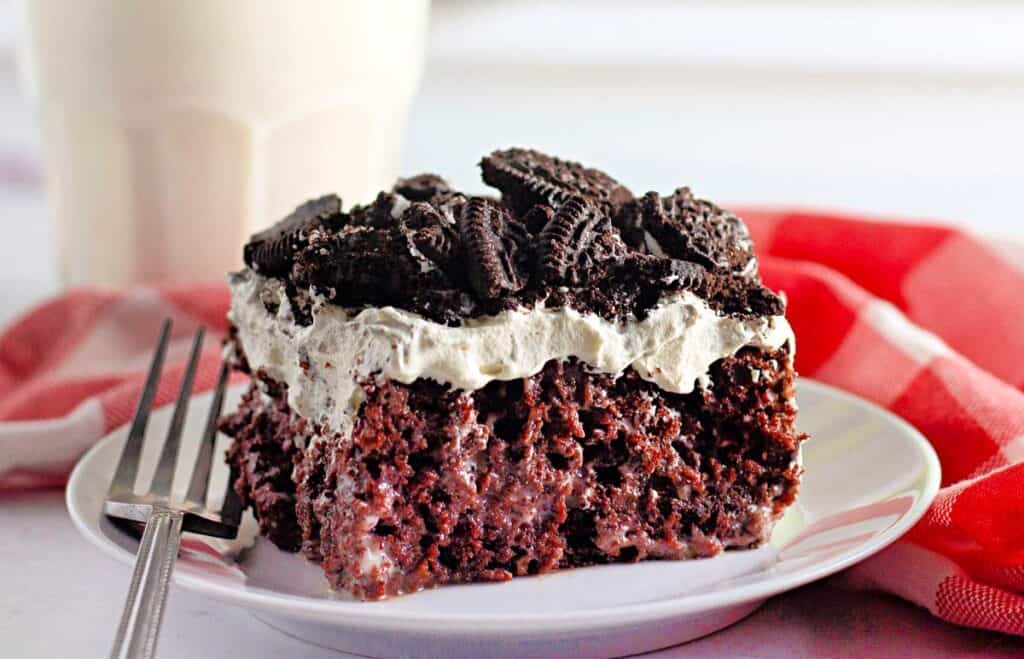 A slice of chocolate cake topped with whipped cream and crushed cookies on a white plate, with a fork and a red checkered cloth in the background.