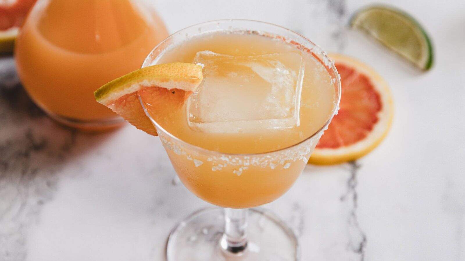 A cocktail glass filled with a light orange drink, garnished with a grapefruit wedge and a large ice cube. The glass has a salted rim and sits on a white surface with a grapefruit slice and a lime wedge in the background.