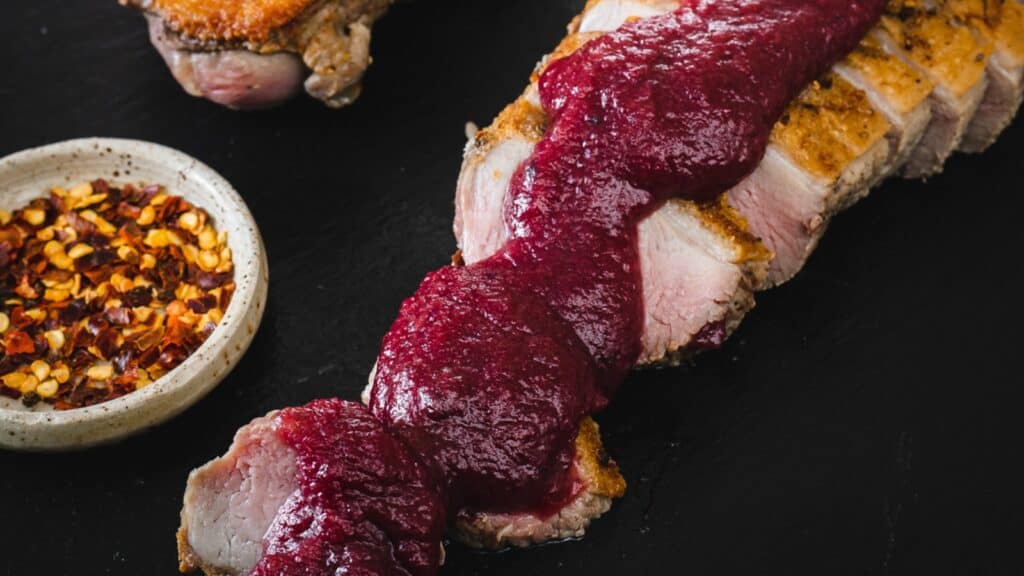 Sliced duck breast with a dark sauce is served on a black slate. Accompanied by a side of kale salad and a small bowl of red pepper flakes. The wooden table background adds a rustic touch.
