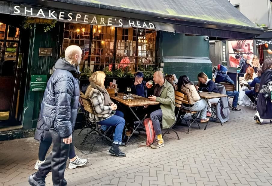 People sit at outdoor tables eating and drinking in front of Shakespeare's Head pub on a city street, enjoying their Pub-cations; a man walks by in the foreground.