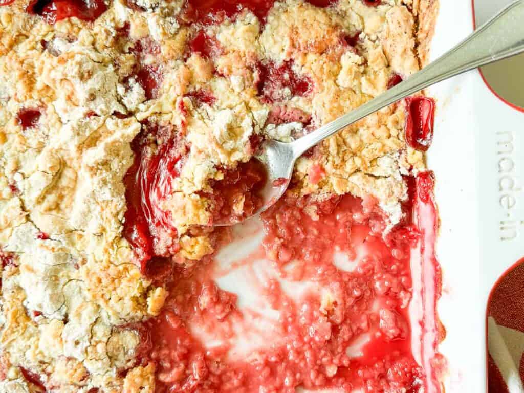 A baking dish with a serving of cherry cobbler removed, revealing the red, juicy filling and golden, crumbly topping. A spoon rests in the dish.