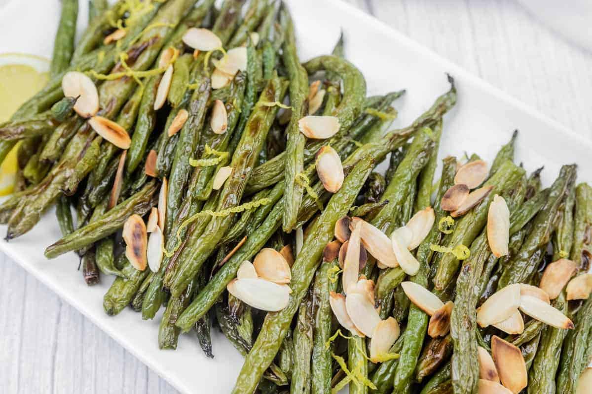 A rectangular white plate with roasted green beans topped with sliced almonds and lemon zest.