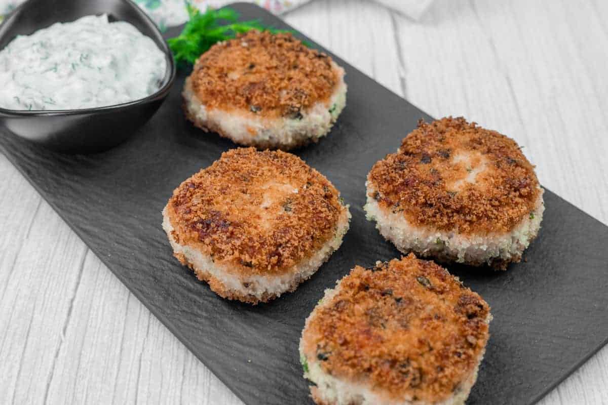 Four Salmon Cakes are arranged on a black slate serving board, accompanied by a small bowl of white sauce with herbs.