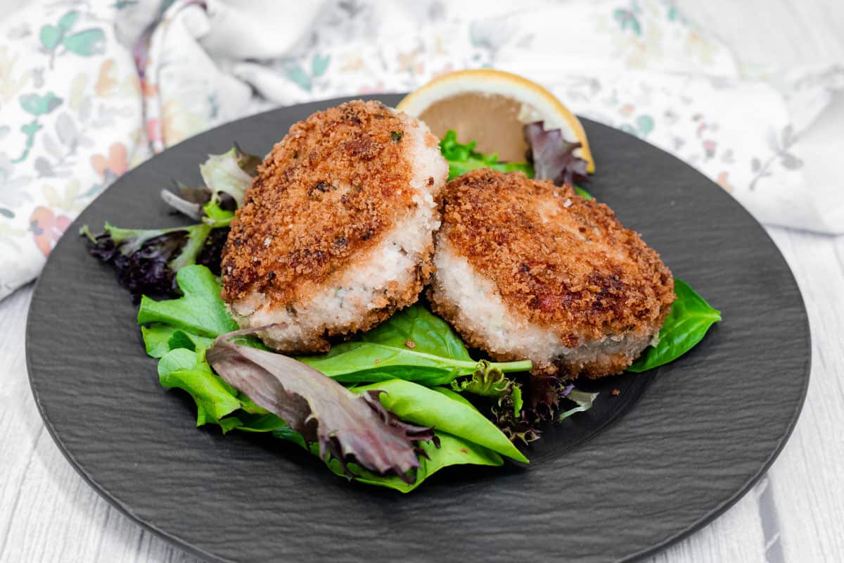 Two Salmon Cakes rest on a bed of mixed greens with a lemon wedge on a black plate, set against a light wood background.