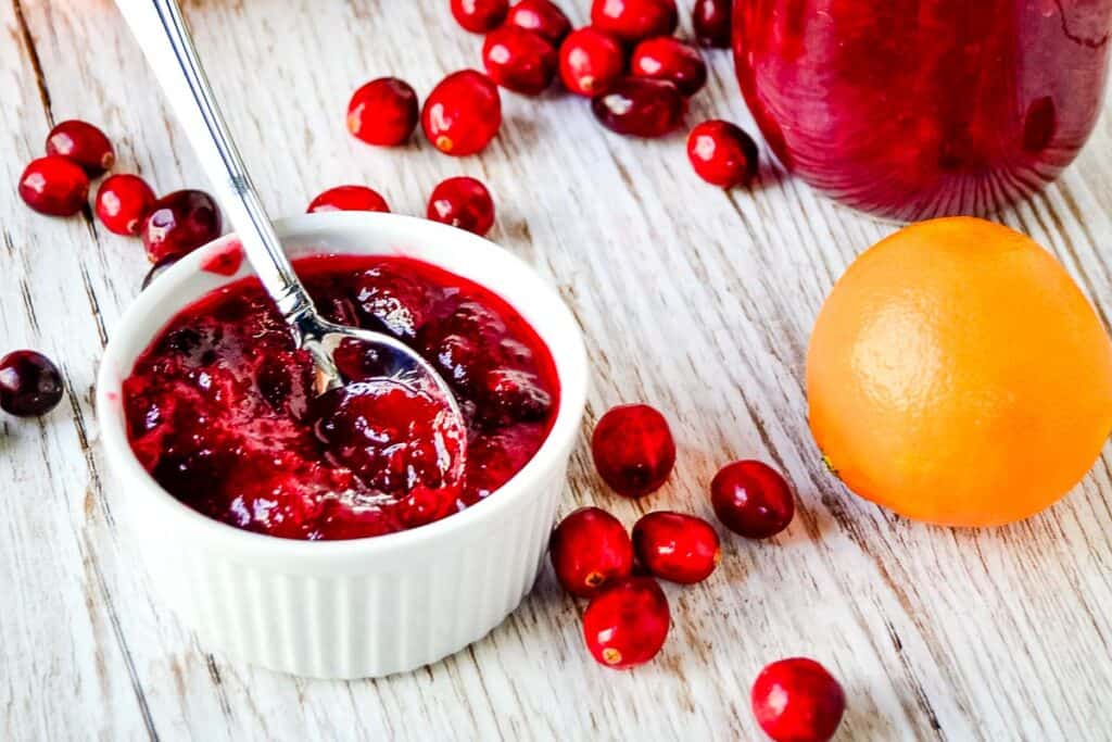 A bowl of cranberry sauce with a spoon, surrounded by fresh cranberries and an orange, on a wooden surface.