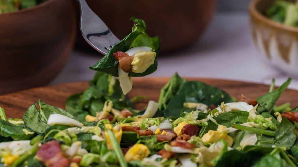 A fork holds a bite of spinach salad with chopped eggs, bacon, and cheese over a wooden bowl filled with the same salad. Two blurred bowls are in the background.