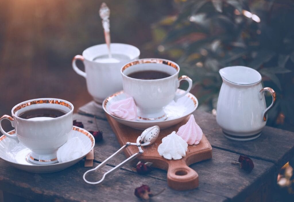 Two cups of coffee on saucers, a sugar bowl, a creamer, and meringues sit charmingly on a wooden table outdoors, evoking the cozy aesthetics of a cottagecore kitchen.