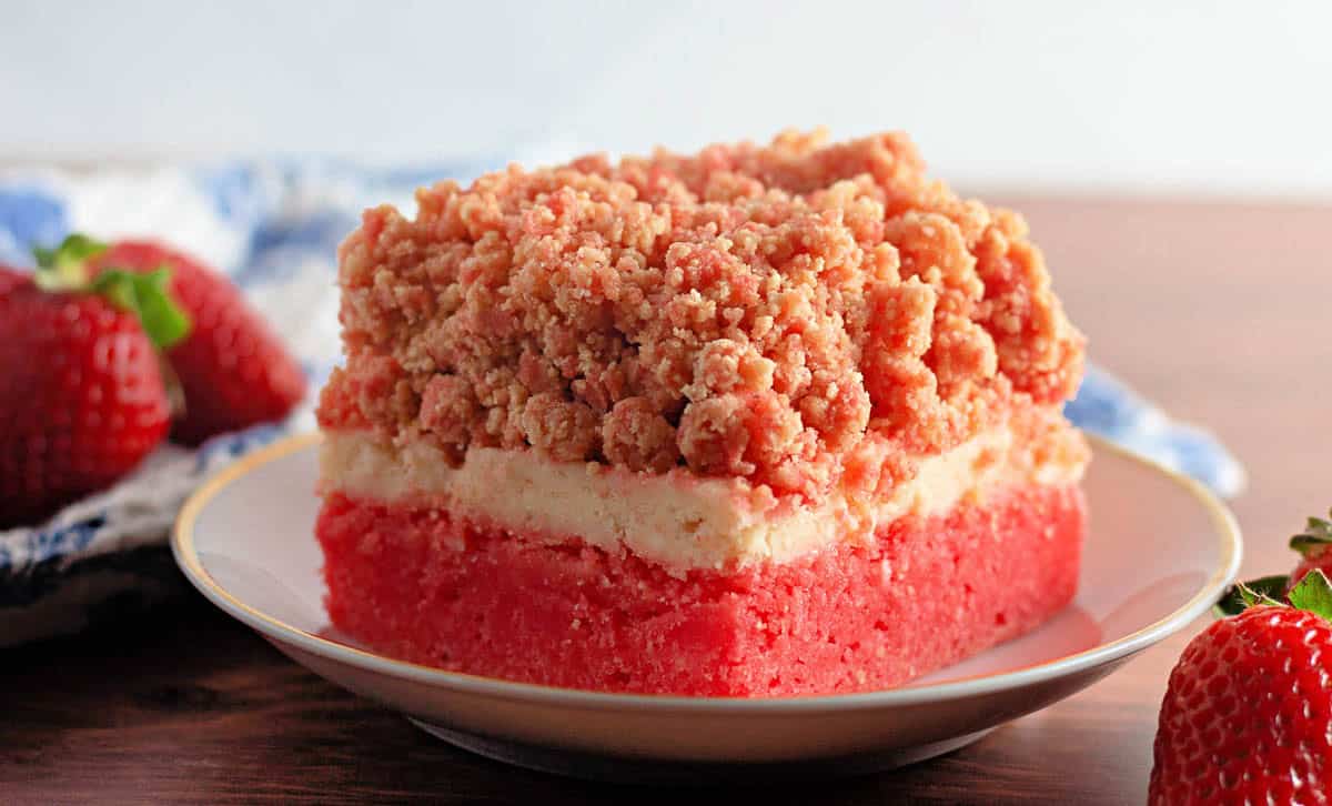 A slice of strawberry crumb cake graces a white plate, embraced by fresh strawberries and a blue-patterned cloth. It captures the essence of delightful strawberry crunch bars on a rustic wooden surface.