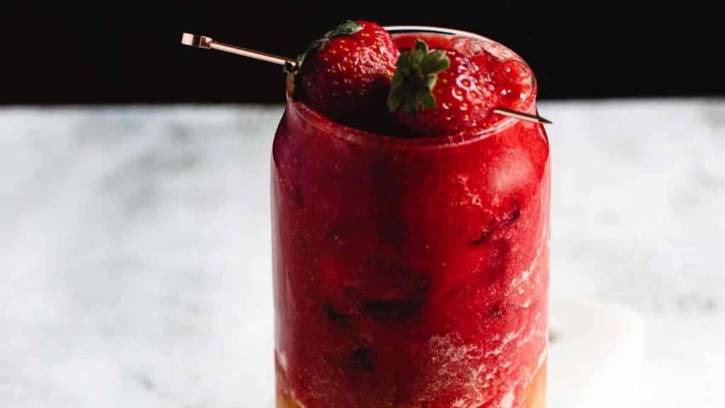 A glass filled with red and yellow smoothie, topped with whole strawberries and a skewer, on a marbled surface.