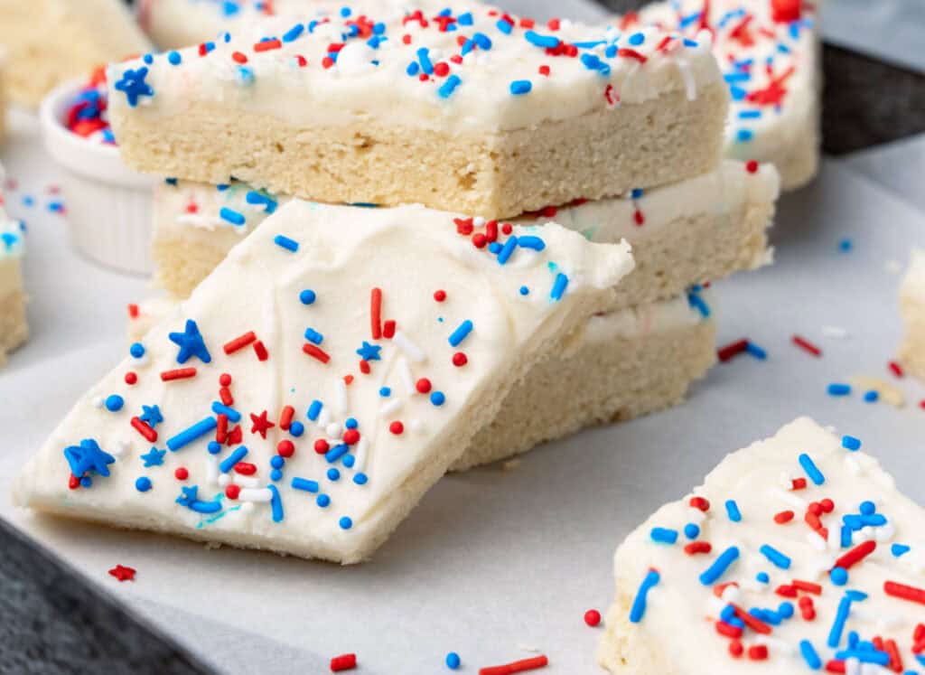 Frosted sugar cookie bars stacked on parchment paper, topped with white icing and red, white, and blue sprinkles.