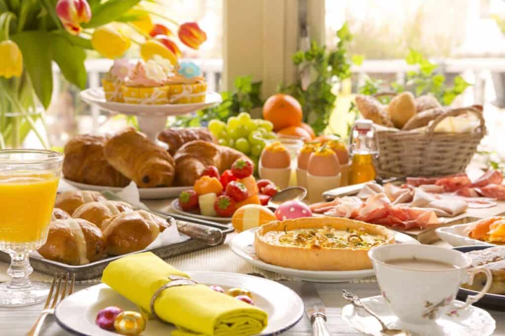 Amidst a delightful breakfast spread with pastries, fruit, quiche, meats, eggs, juice, and coffee on the table, tulips flourish by the window overlooking lush greenery. While fancy Easter dinners are dying out in popularity, this morning gathering thrives with simple elegance.