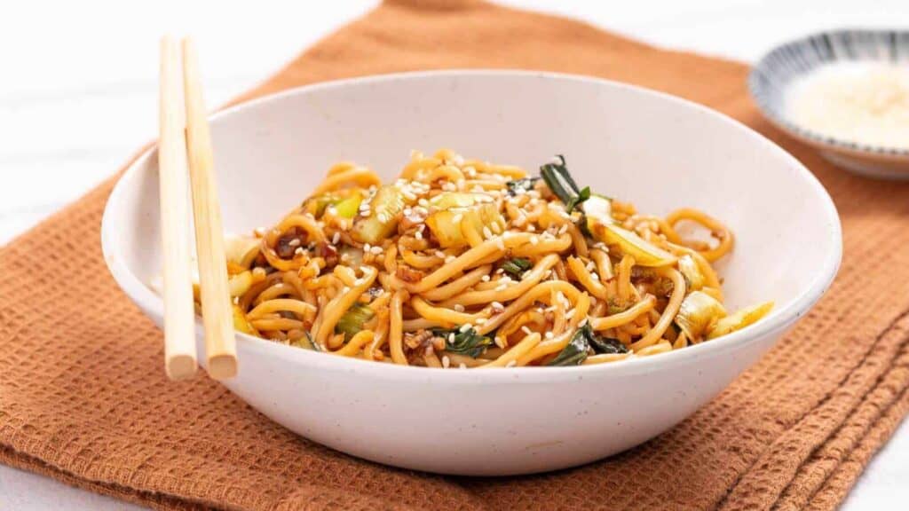A white bowl of stir-fried noodles with vegetables and sesame seeds, placed on a brown textured cloth, with chopsticks resting on the bowl.