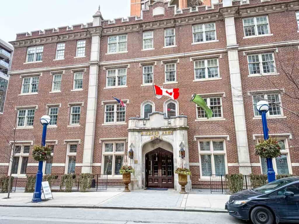 A brick building with three flags above the entrance, including a Canadian flag, and a sign reading &ldquo;University Club of Toronto&rdquo; on a city street.