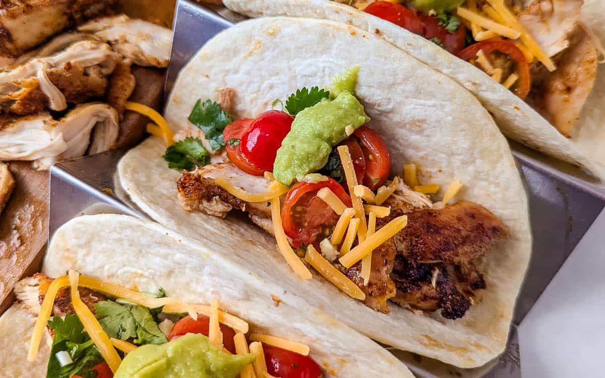 Close-up of grilled chicken tacos topped with shredded cheese, cherry tomatoes, guacamole, and cilantro, served in flour tortillas.