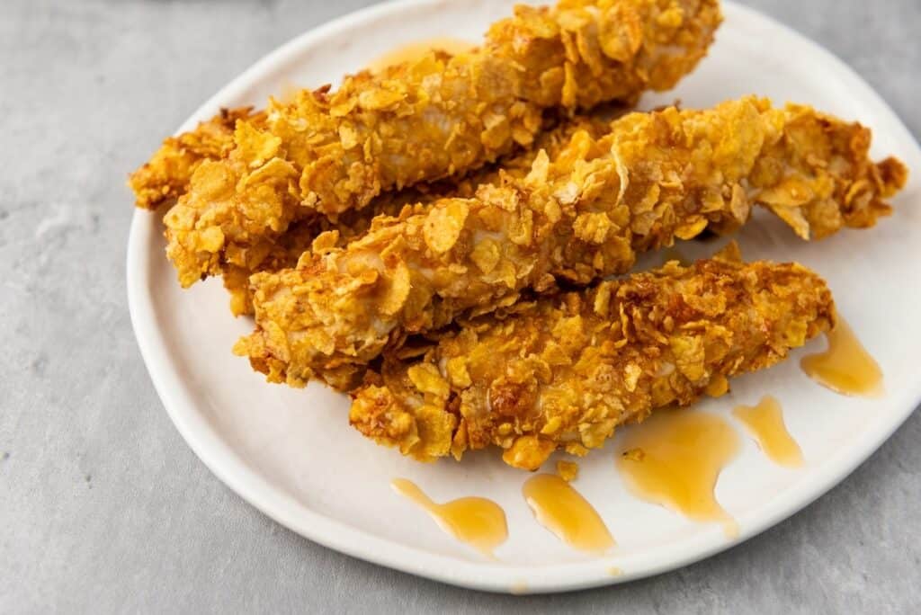 Three crispy, golden-brown breaded chicken tenders are arranged on a white plate with a drizzle of sauce beside them.