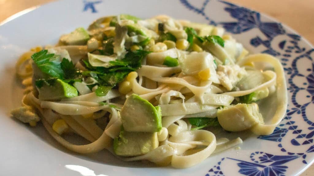 A plate of pasta with avocado, corn, and greens on a white plate with a blue floral pattern.