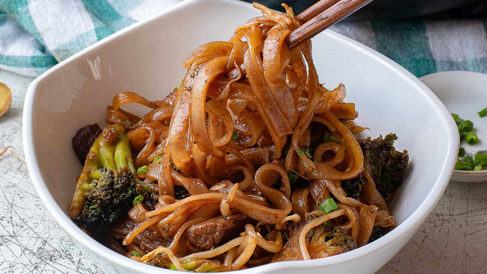 Two bowls of beef chow fun stir-fried with broccoli and bean sprouts, garnished with sesame seeds. The dishes are served in gray bowls, and the background shows another bowl with ingredients. A pair of chopsticks is placed beside the front bowl.
