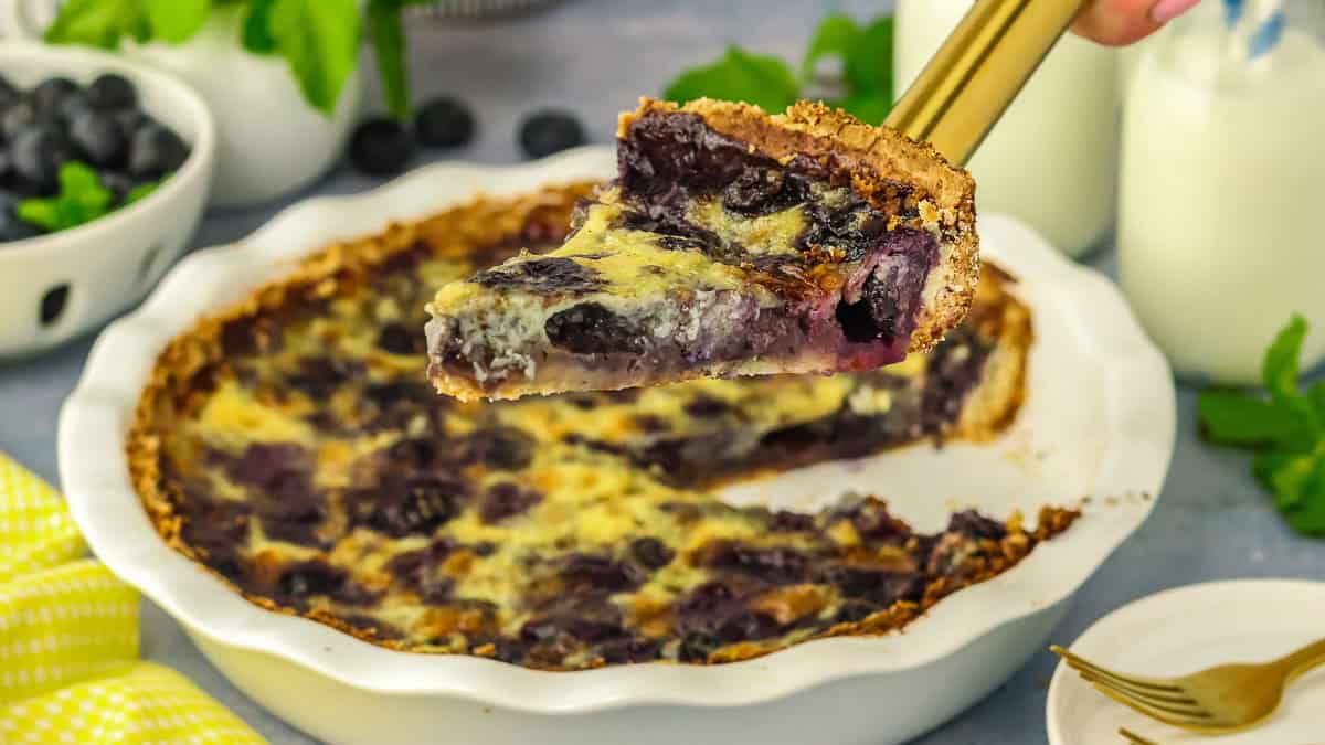 A slice of blueberry pie with a crumbly crust being lifted from a pie dish, surrounded by fresh blueberries and milk bottles.
