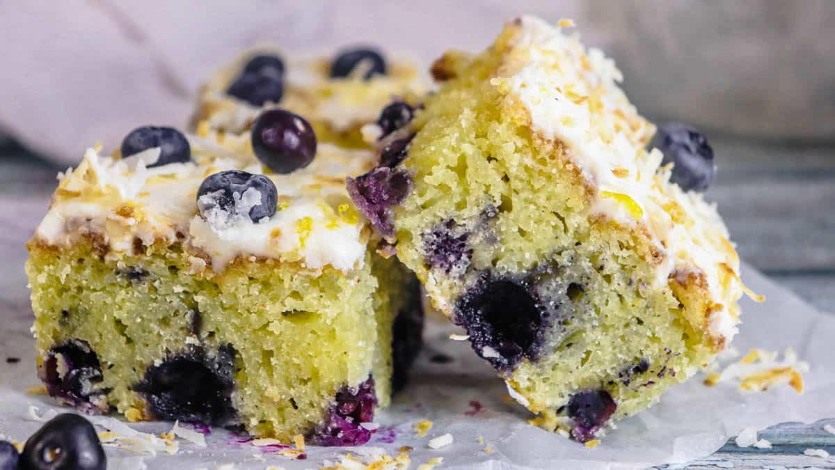 Two slices of blueberry cake topped with frosting, shredded coconut, and fresh blueberries on parchment paper.
