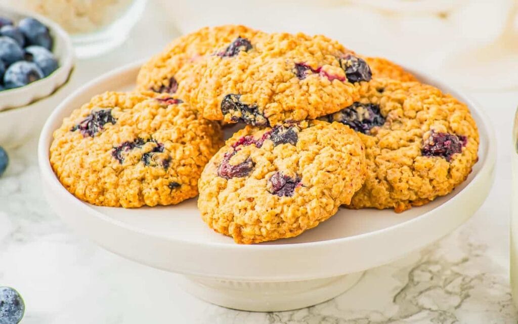 A white cake stand holds a stack of oatmeal cookies with visible blueberries. A bowl of fresh blueberries sits nearby on a marble surface.