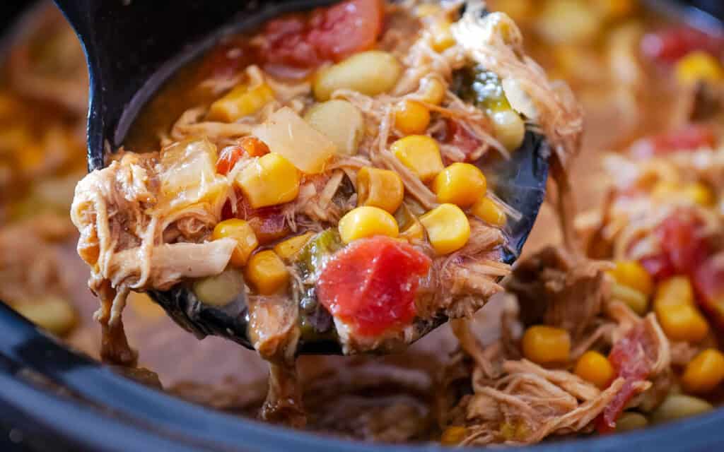 A close-up of a ladle lifting a stew with shredded meat, corn, lima beans, diced tomatoes, onions, and broth.