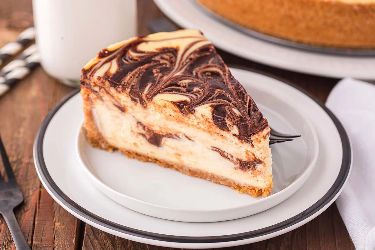 A slice of marble cheesecake with a chocolate swirl pattern, served on a white plate with a black rim.