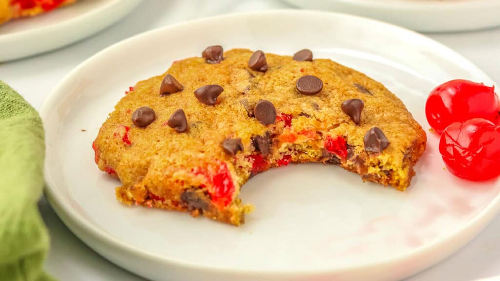 A cookie with chocolate chips and cherries on a white plate, partially eaten, with two whole cherries beside it.