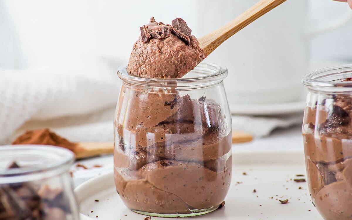 A wooden spoon lifts a portion of chocolate mousse with chocolate shavings from a small glass jar, surrounded by other jars on a white tray.