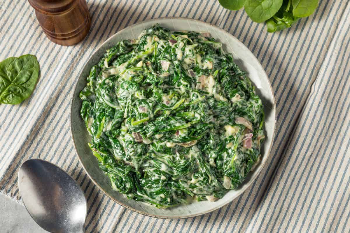 A bowl of creamed spinach on a blue striped cloth, with a spoon and fresh spinach leaves nearby.