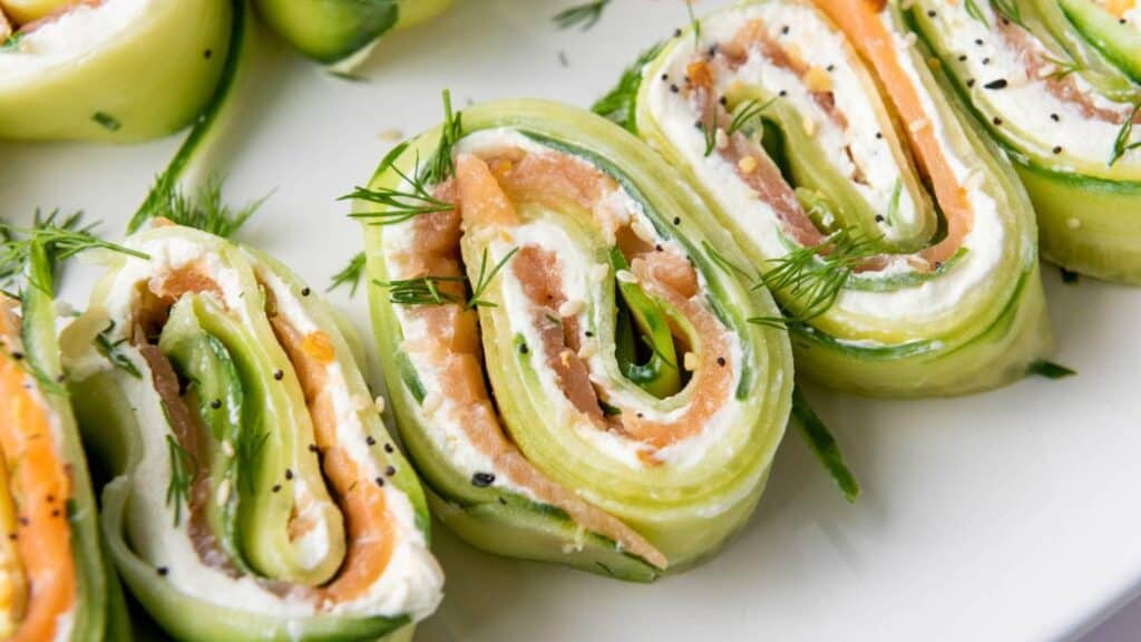 Close-up of cucumber rolls filled with cream cheese, smoked salmon, and garnished with dill on a white plate.