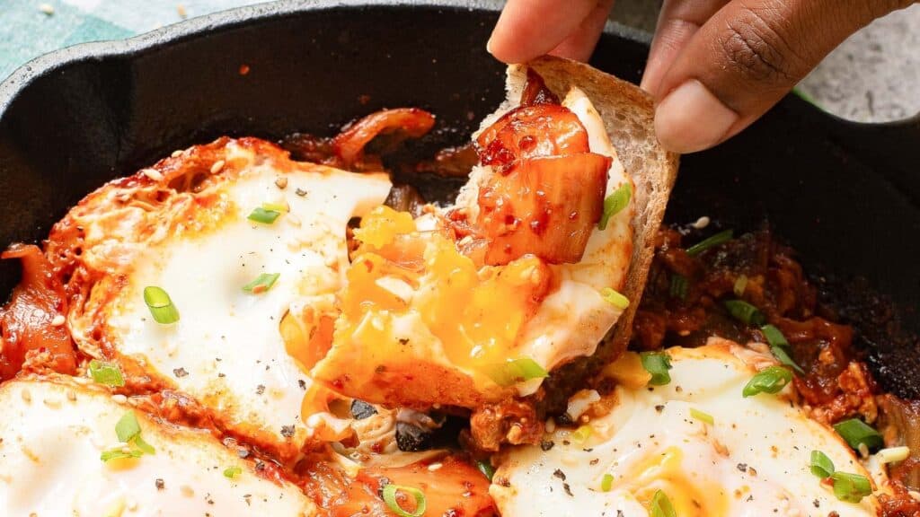 A hand dips a slice of bread into a skillet dish with eggs cooked in a tomato-based sauce, garnished with chopped green onions.