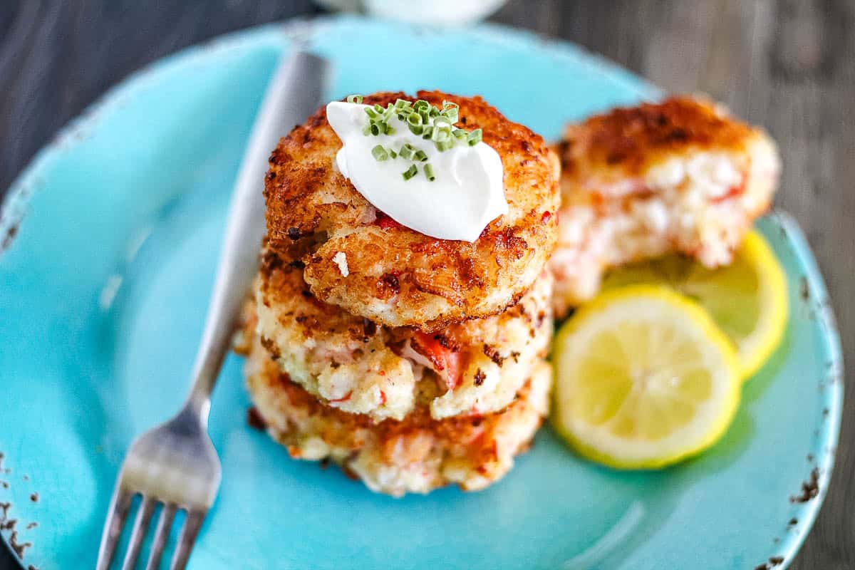 Three crab cakes stacked on a blue plate, topped with sour cream and chopped chives, with lemon slices and a fork on the side.