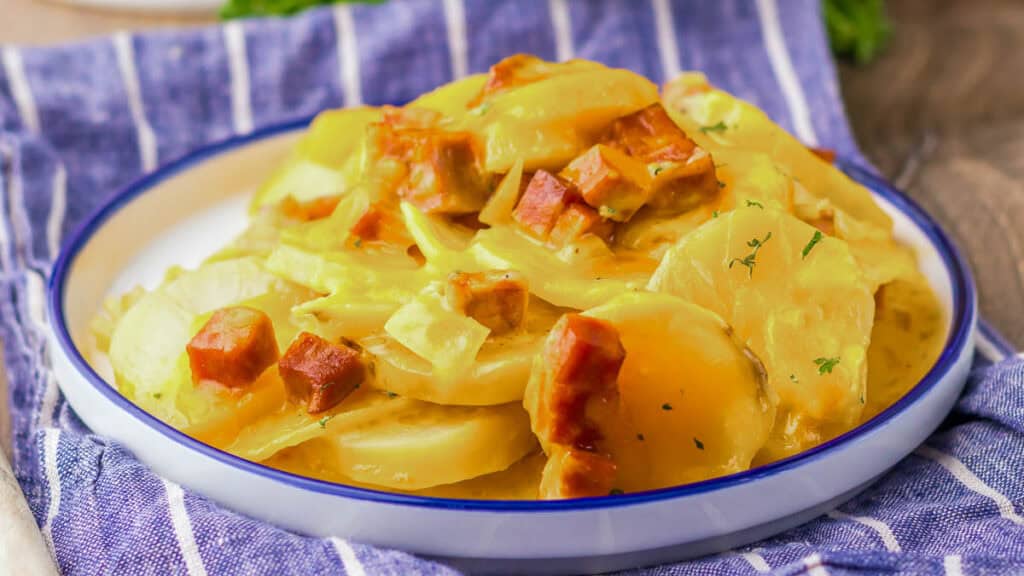 A plate of scalloped potatoes with chunks of ham, topped with a creamy sauce and garnished with herbs, placed on a blue-striped cloth.