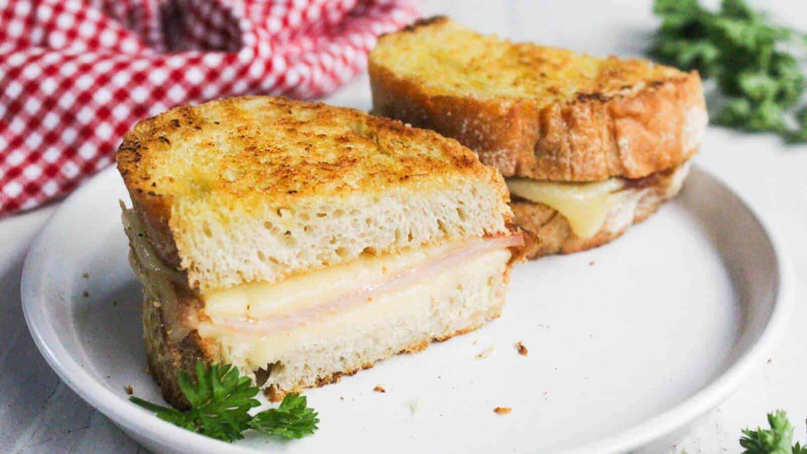 A grilled cheese sandwich with ham is halved and placed on a white plate. A red checkered cloth and green herbs are in the background.
