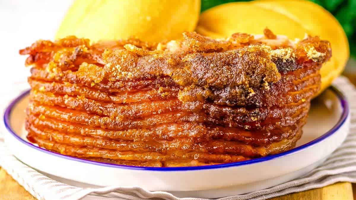 Close-up of a honey-glazed ham with caramelized edges on a plate, accompanied by two bread rolls in the background.