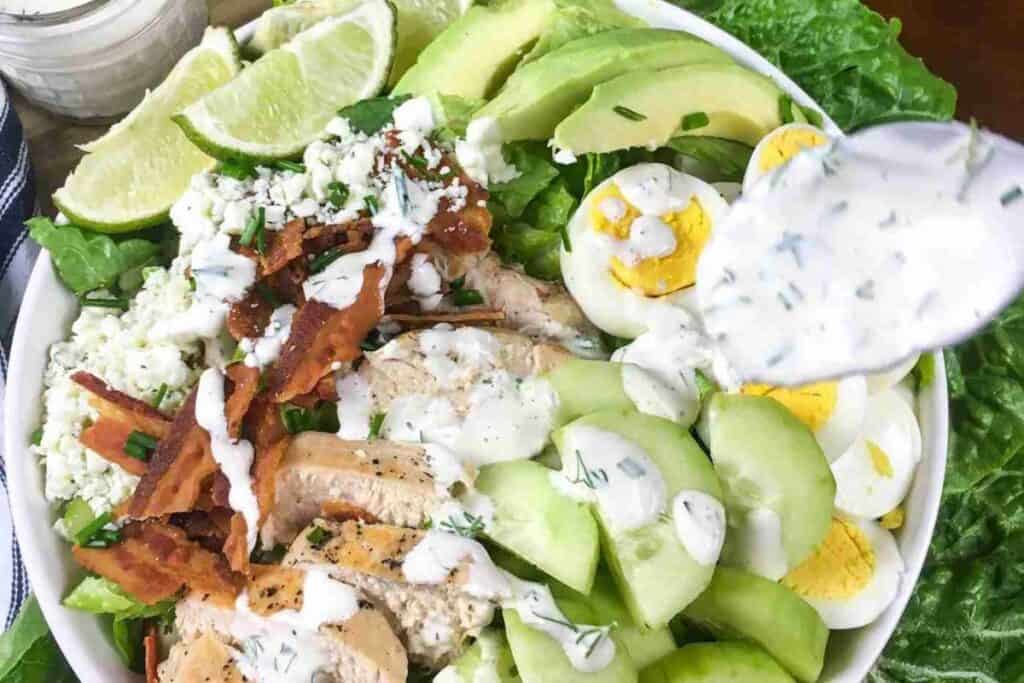 A bowl of salad with sliced grilled chicken, bacon, egg, avocado, cucumber, cheese, lime wedges, and ranch dressing being drizzled on top.
