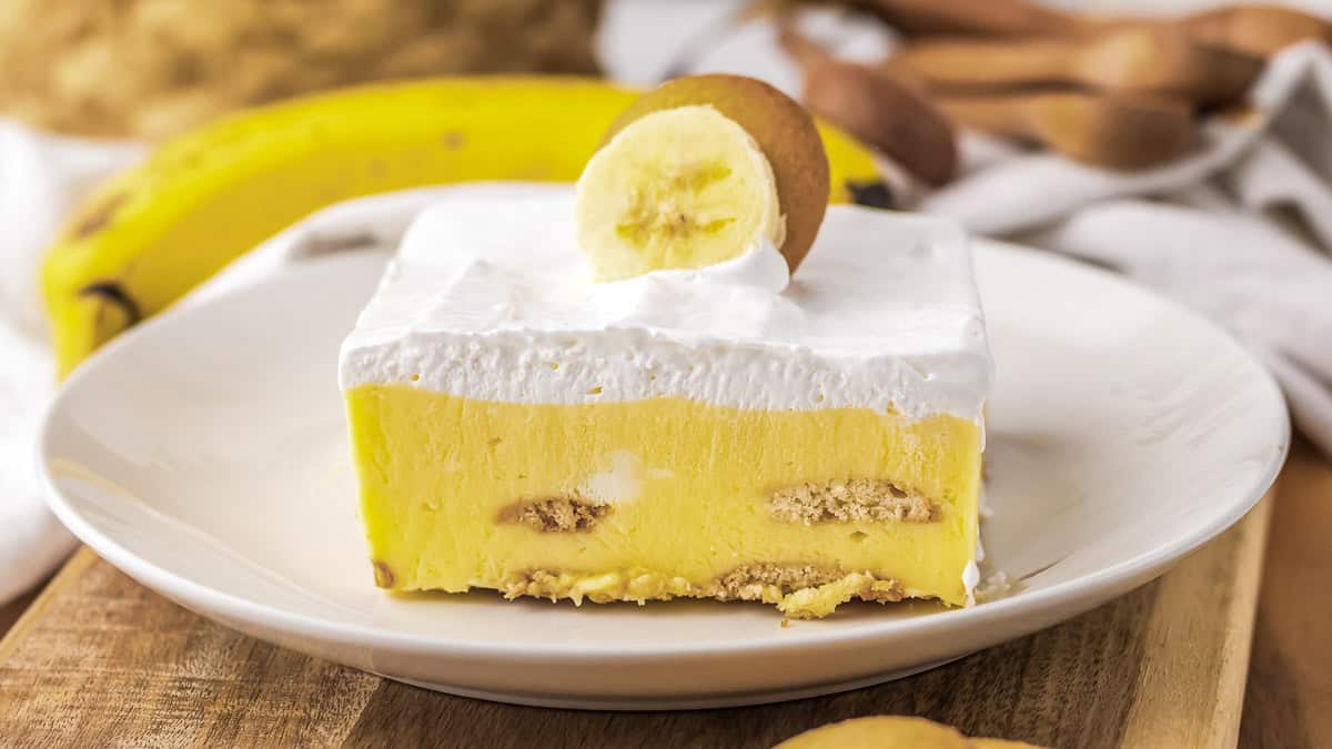 A slice of banana pudding with layers of vanilla pudding, whipped cream, and cookies on a white plate. A banana slice and cookie garnish the dessert. A banana and utensils in the background.