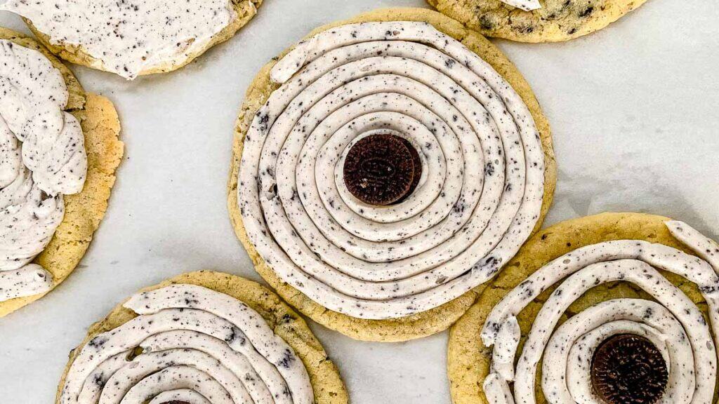 Cookies topped with spirals of white and speckled cream, centered with a chocolate cookie piece, arranged on a white surface.