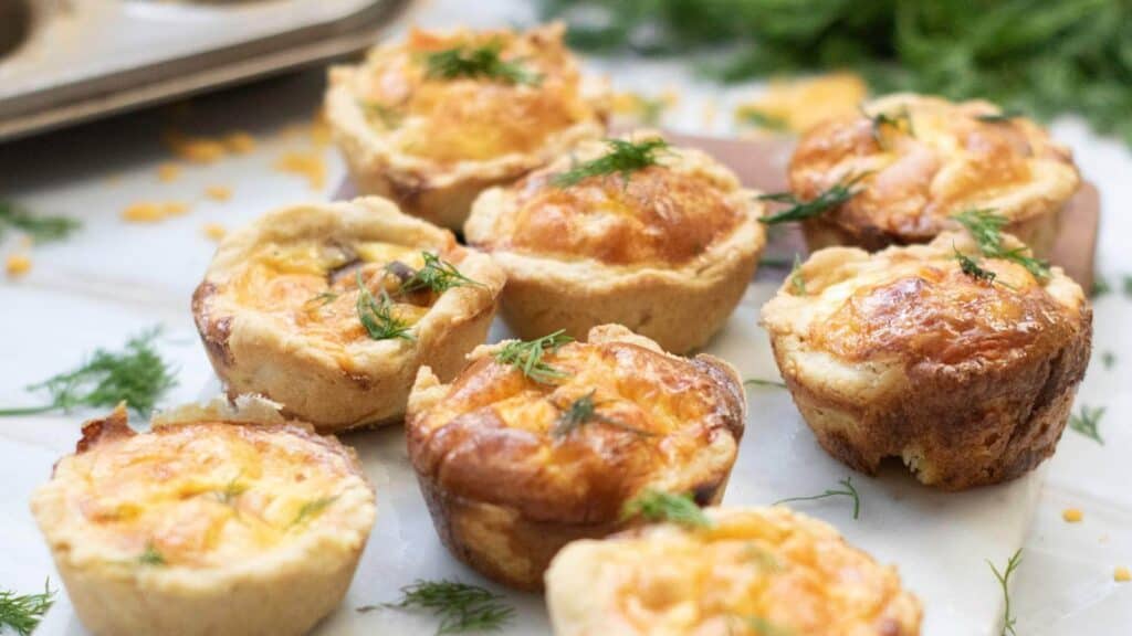 Mini quiches with golden tops are arranged on a marble surface, garnished with fresh dill.