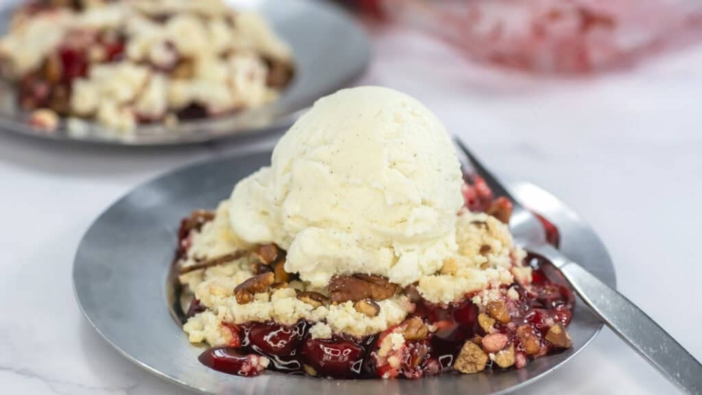 A dessert plate with cherry crumble topped with a scoop of vanilla ice cream and pecans, placed on a shiny plate with a spoon beside it. Another plate is slightly blurred in the background.