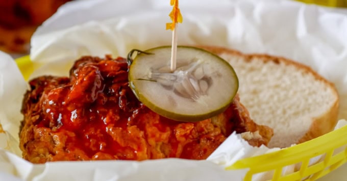 A piece of fried chicken with sauce is topped with a pickle slice and served on a sandwich bun in a yellow basket with white paper.