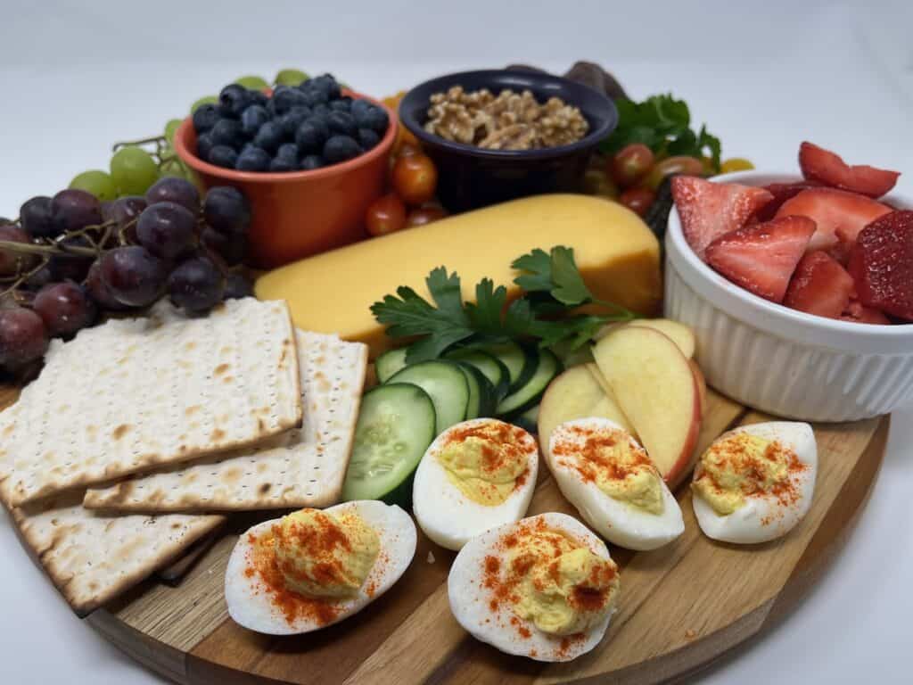 A wooden platter with deviled eggs, sliced cucumber, apple, strawberries, blueberries, grapes, walnuts, cheese, parsley, cherry tomatoes, and crackers.